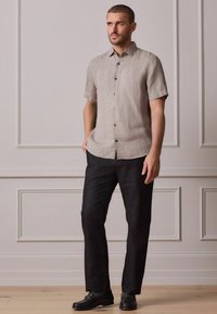 Kurzärmeliges, hellbeiges Leinenhemd mit einem Kragen und Knöpfen, kombiniert mit schwarzen Hosen und schwarzen Loafers, stehend vor einem neutralen Hintergrund.