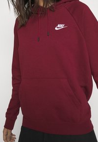 Burgundy Nike-hoodie med framficka, justerbar dragsko och vitt logo. Mjuk textur, lång ärm och ribbad mudd och nederkant.