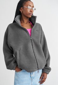 Felpa grigia con zip e colletto alto, tasche laterali e logo ricamato. Indossata sopra una maglietta rosa. Tessuto morbido e vestibilità comoda.