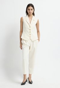Ivory mouwloos vest met vijf gouden knopen en een puntige zoom, gecombineerd met ivoorkleurige, tapered broek en zwarte hoge hakken.