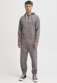 Gray cotton zippered onesie with a hood, featuring drawstrings, elastic cuffs, and a small logo patch. Worn with black high-top sneakers.