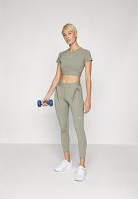 Oliven grønn cropped treningsgenser med Nike-logo, sammen med matchende høyt satte tights med subtile teksturdetaljer og en blå hantel.