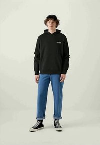 Giovane che indossa un hoodie nero, jeans blu arrotolati, sneakers nere alte e un cappellino nero, in piedi su uno sfondo semplice.