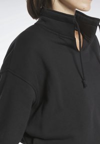 Žena oblečená v černém svetru s vysokým límcem a šňůrkou, snímána od ramen po bradu na světlém pozadí.