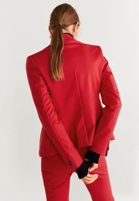Blazer rouge sur mesure avec une texture lisse, un design structuré et des accents noirs sur les manches. Doté d'une fente unique à l'arrière et d'un léger rembourrage aux épaules.