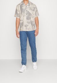 Camisa de manga corta en color beige con un patrón abstracto en gris; combinada con jeans azules y zapatillas blancas, de pie sobre un fondo liso.