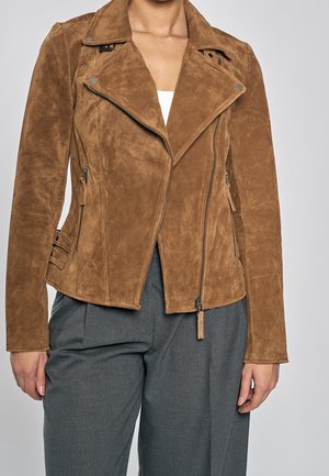 Bruine suède bikerjas met asymmetrische rits, metalen drukknopen, ritssluitingen en gespdetails, gedragen met grijze broek.