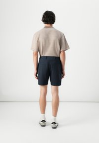 Cămașă scurtă bej cu dungi maro, purtată cu pantaloni scurți bleumarin. Ținuta are o croială lejeră și este completată cu încălțăminte albă și neagră.