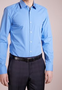 Lichtblauw overhemd met lange mouwen, een puntige kraag en een knoopsluiting aan de voorkant, gecombineerd met donkere, patroonbroeken en een zwarte riem.