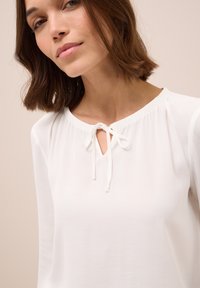 Blusa bianca in tessuto leggero, con apertura a chiave e dettaglio fiocco al collo, dotata di maniche arricciate.
