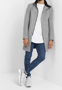 Cappotto di lana grigio con colletto nero, bottoni a pressione e taglio dritto. Abbinato con una maglietta bianca, jeans blu e sneakers grigie.