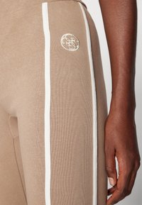 Ljusbruna leggings med vita sidostråk, ribbad textur och en broderad logga på låret. Närbild som visar handdetalj.