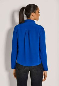 Langärmliges Hemd aus leuchtend blauem Stoff, mit Kragen und plissierter Rückendetail. Kombiniert mit dunklen Jeans für einen kompletten Look.