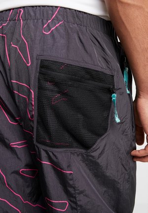 Zbliżenie osoby noszącej ciemne szorty z różowymi wzorami liniowymi oraz czarną siatkową tylną kieszenią na zamek z turkusowym uchwytem.