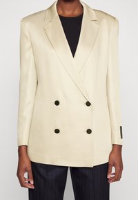 Blazer de doble botonadura en crema con una textura suave, solapas afiladas y botones negros. Combinado con una blusa negra y pantalones de rayas.
