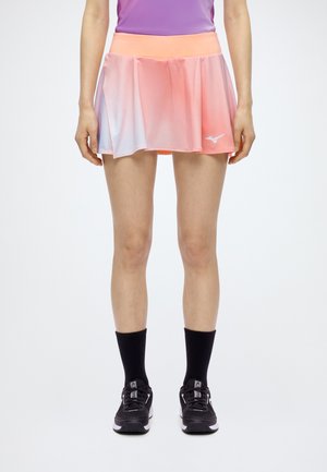 Modèle portant une jupe de sport plissée rose corail, un haut violet, des chaussettes noires et des baskets noires.