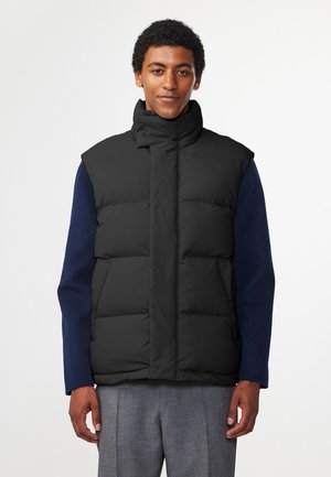 Zwarte gewatteerde vest met hoge kraag, ritssluiting aan de voorkant en meerdere grote gestikte secties; gedragen over een marineblauwe longsleeve shirt.