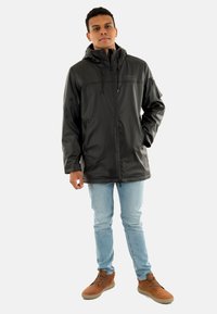 Giacca nera impermeabile con cappuccio, chiusura con zip e cordini regolabili, abbinata a jeans azzurri chiari e stivali marroni.