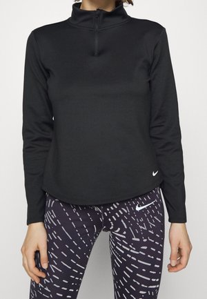 Vrouw draagt een zwart top met lange mouwen en een halve rits, en zwarte sportleggings met witte abstracte patronen en Nike-logo's.