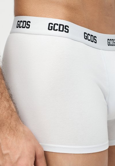 Gros plan d'une personne portant un boxer blanc avec une ceinture blanche portant l'inscription "GCDS" en lettres noires.