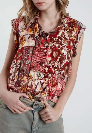 Donna che indossa una blusa patchwork floreale senza maniche nei toni del rosso, marrone e arancione, che sistema un bottone sui jeans azzurri.
