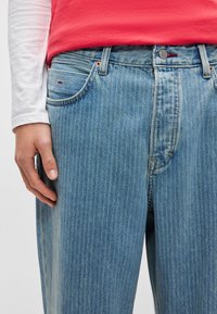 Φωτεινό γαλάζιο τζιν με κάθετες ρίγες, πλαϊνές τσέπες και κλείσιμο με κουμπί. Χέρι ακουμπάει στον μηρό. Συνδυασμός βαμβακιού, casual σχεδίαση.