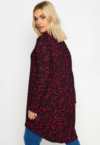 Svart tunika med röd blommönster, långa ärmar och en längre bakdesign. Mjukt, texturerat material, avslappnad passform, lämplig för vardagsbruk.