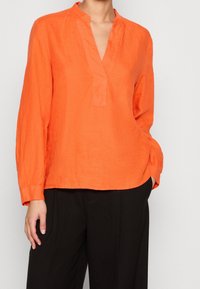 Chemisier en lin orange avec un col mandarin, des manches longues, une patte de boutonnage half-placket et une coupe décontractée. Associé à un pantalon noir.