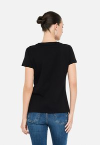 Kvindeligt ansigt med m�ørkt hår i en knold iført sølv hængende øreringe, en simpel sort T-shirt med korte ærmer og blå denim bukser, vendt væk.