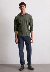 Polo à manches longues vert olive avec un col, associé à un pantalon droit bleu marine. Le modèle porte des chaussures noires, debout sur une surface texturée.