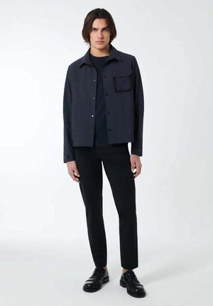 Giacca blu navale con bottoni, dotata di taschino sul petto, abbinata a una maglietta nera, pantaloni neri e scarpe nere. Design elegante e minimalista, texture liscia.