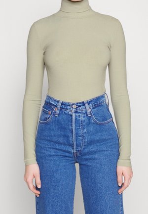 Personne portant un haut vert clair côtelé à col roulé et manches longues, rentré dans un jean en denim bleu taille haute, sur un fond uni.