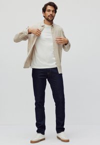 Lichtbeige overhemd met knopen over een wit shirt, gecombineerd met donkerblauwe spijkerbroek en witte sneakers met een tan zool. Minimalistisch ontwerp.