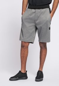 Grå träningsshorts med ett elastiskt svart midjeband, sidofickor, slät textur och en liten logotypapplikation på vänster ben. Matchas med svarta sneakers.