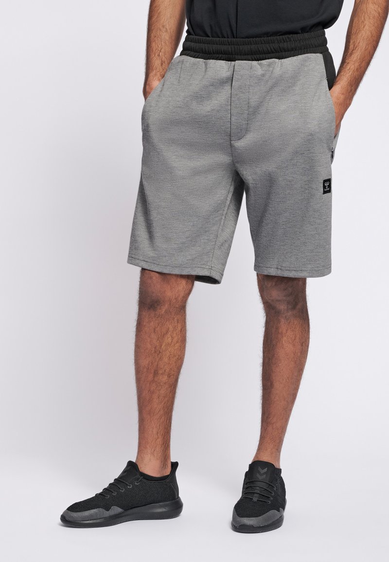 Grå träningsshorts med ett elastiskt svart midjeband, sidofickor, slät textur och en liten logotypapplikation på vänster ben. Matchas med svarta sneakers.