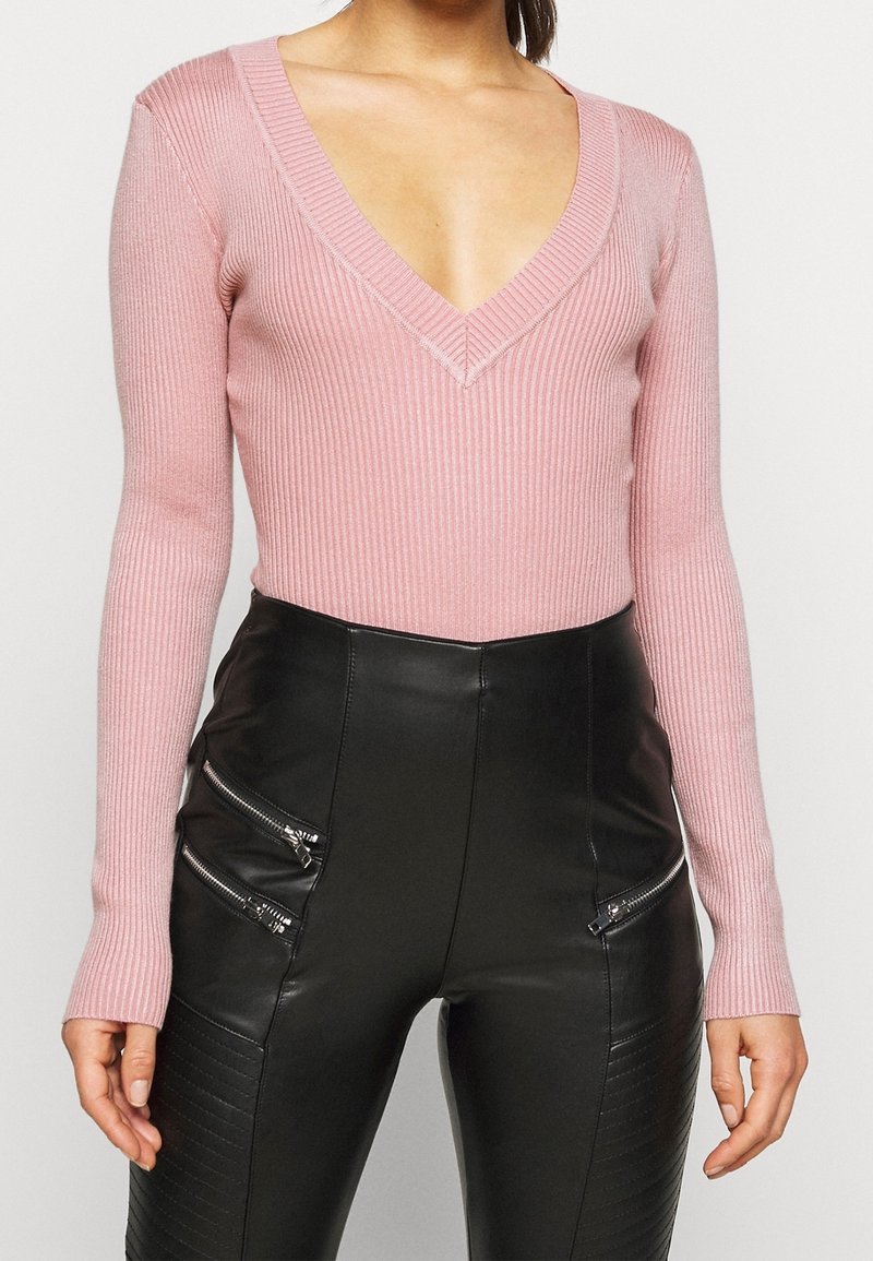 Ljusrosa ribbad långärmad body med djup V-ringning, kombinerad med svarta byxor i fuskläder med dragkedjeförsedda detaljer.