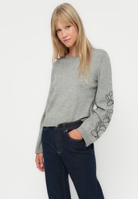 Grå strikkegenser med rund hals og lange ermer, prydet med svart blomsterbroderi på venstre erme. Kombinert med mørke denimjeans.