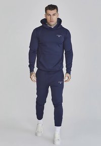 Marineblauwe hooded trui en bijpassende joggingbroek van zachte stof. Voorzien van een logo op de borst en een buidelzak op de hoodie.