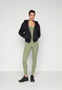 Sweatshirt preta com fecho de correr e dois bolsos, usada sobre um body verde-oliva justo e leggings combinando. Ténis desportivos pretos completam o conjunto.