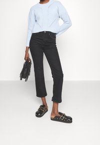 Maglione azzurro chiaro lavorato a maglia con motivo testurizzato; jeans neri a vita alta con orlo grezzo; zoccoli neri con dettagli in oro e borchie; borsa con frange.