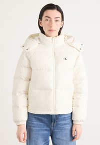 Puffjakke i off-white farge med høy krage, frontglidelås, elastiske mansjetter og logo på brystet. Glatt, polstret tekstur.