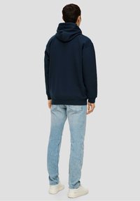 Hombre vestido con una sudadera azul oscura, jeans azul claro y zapatillas blancas, de pie girado de espaldas contra un fondo gris claro liso.