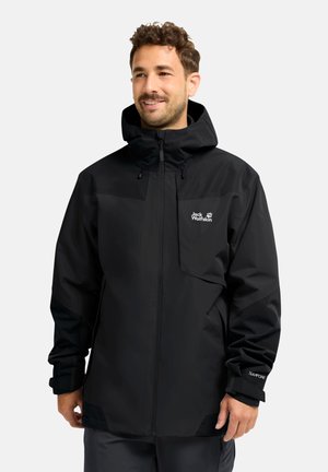Mann trägt eine schwarze wasserdichte Jack-Wolfskin-Kapuzenjacke mit Frontreißverschluss und Logo auf der Brust, steht vor einem einfarbigen hellen Hintergrund.