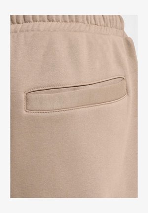 Pantalonii de trening bej au o textură netedă și un elastic la talie, cu un detaliu de buzunar orizontal integrat în spate.