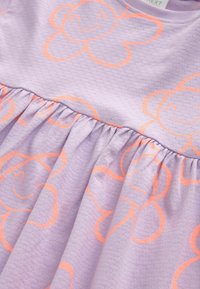 Tissu violet clair avec des motifs de fleurs souriantes orange et un détail de couture froncée.