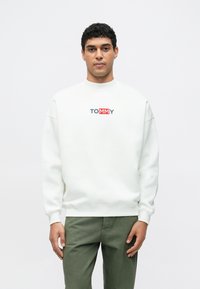 Bílá mikina s kulatým výstřihem a žebrovanými manžetami, na hrudi je barevné logo "TOMMY". Model má na sobě zelené kalhoty.