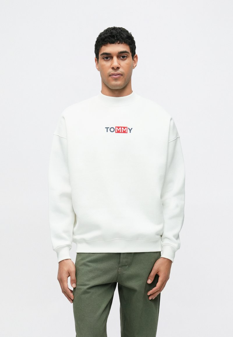 Bílá mikina s kulatým výstřihem a žebrovanými manžetami, na hrudi je barevné logo "TOMMY". Model má na sobě zelené kalhoty.