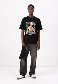 Sort t-shirt med et trykt grafisk motiv af en karakter, parret med løse grå jeans og hvide sneakers. En lille sort taske holdes i den ene hånd.