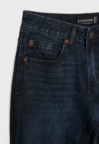 Jean en denim bleu foncé avec poche avant, rivets en cuivre, taille avec bouton et étiquette de la marque Stradivarius à l'intérieur de la taille.
