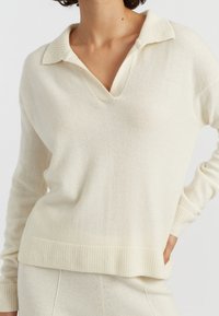 Hellbeige Pullover mit V-Ausschnitt und geripptem Kragen. Weiche Textur, lange Ärmel und gerippter Saum. Kombination mit passender Hose.